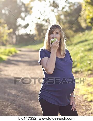 Stock Image - Pregnant Caucasian woman eating apple outdoors. Fotosearch