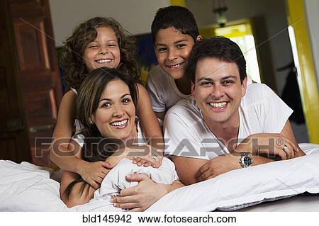 Stock Image - Smiling Hispanic family laying on bed together. Fotosearch