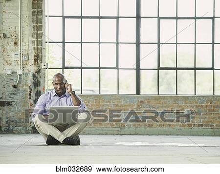 Stock Photo - Black businessman using laptop and cell phone on warehouse floor. Fotosearch