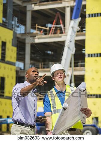Stock Image - Businessman and construction worker reading blueprints at construction site. Fotosearch