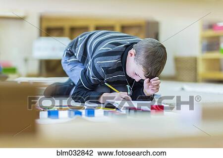 Stock Image - Caucasian boy writing in notebook on floor in classroom. Fotosearch