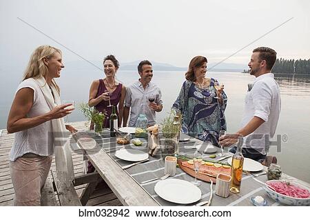 Stock Image - Caucasian friends talking at picnic table at lake. Fotosearch