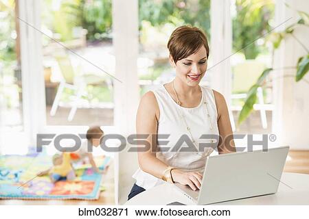 Stock Image - Caucasian mother using laptop at counter. Fotosearch