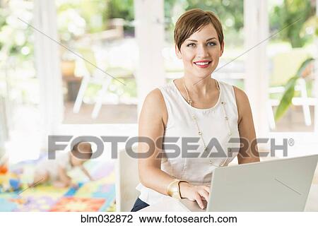 Stock Image - Caucasian mother using laptop at counter. Fotosearch