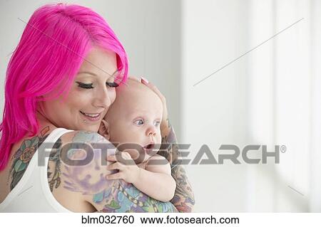 Stock Image - Caucasian mother with pink hair and tattoos cradling baby. Fotosearch