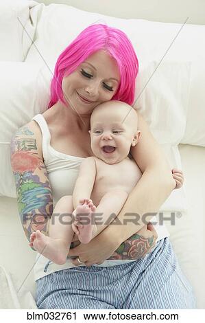 Stock Image - Caucasian mother with pink hair and tattoos cradling baby. Fotosearch