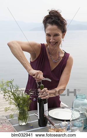 Stock Image - Caucasian woman opening wine bottle at table near lake. Fotosearch
