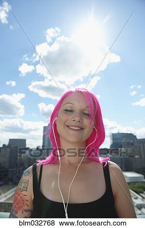 Stock Photo - Caucasian woman with pink hair and tattoos listening to earbuds in city. Fotosearch