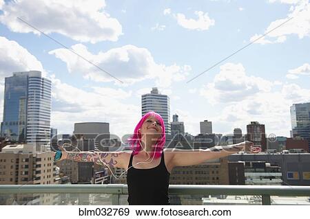 Stock Photo - Caucasian woman with pink hair and tattoos listening to earbuds on urban rooftop. Fotosearch