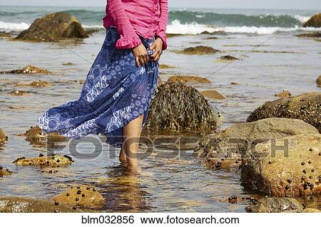 Stock Photograph - Mixed race woman walking in water on rocky beach. Fotosearch