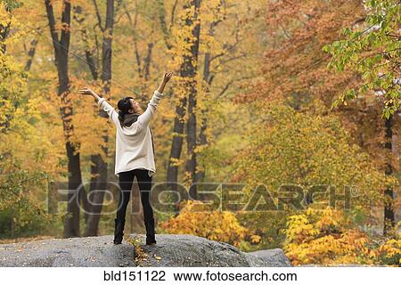Stock Image - Asian woman cheering with arms outstretched on rock in park. Fotosearch