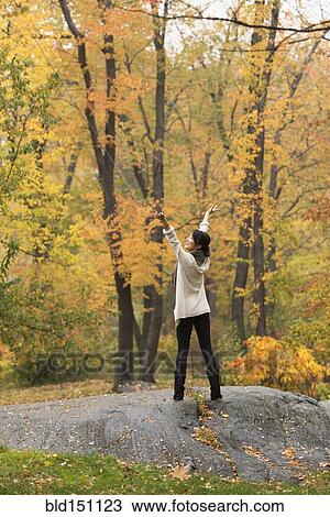 Stock Image - Asian woman cheering with arms outstretched on rock in park. Fotosearch