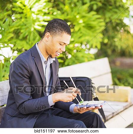 Stock Photo - Black businessman eating sushi on park bench. Fotosearch