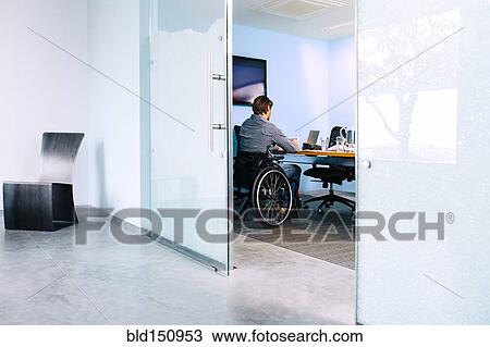 Stock Image - Businessman in wheelchair working at desk in office. Fotosearch