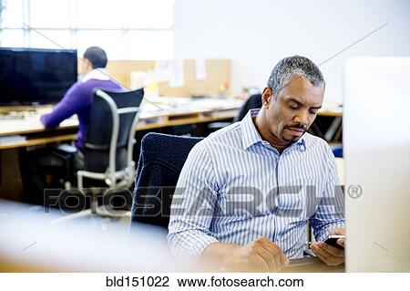 Stock Image - Businessman using cell phone at office desk. Fotosearch
