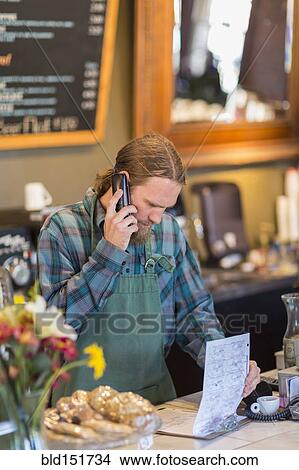 Picture - Caucasian server on telephone reading clipboard in cafe. Fotosearch