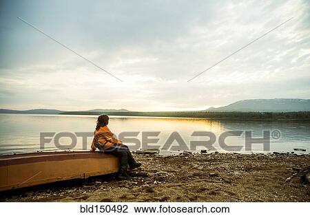 Stock Image - Caucasian woman sitting on dilapidated boat at rural lake. Fotosearch