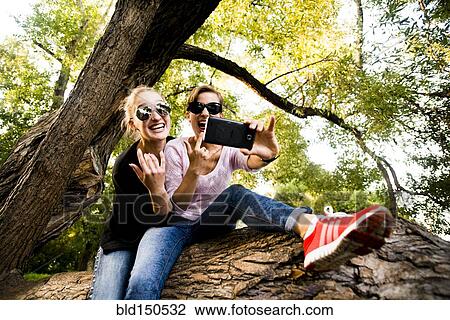 Stock Image - Caucasian women taking cell phone selfie in tree. Fotosearch