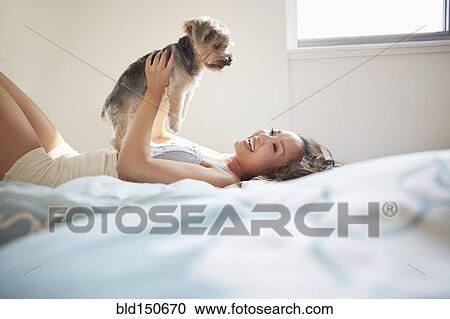 Stock Image - Chinese woman playing with dog on bed. Fotosearch