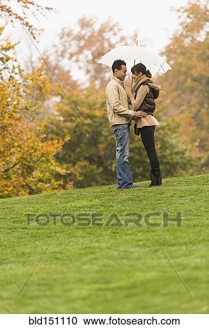 Stock Image - Couple standing with umbrella in park. Fotosearch