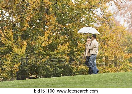 Stock Image - Couple walking with umbrella in park. Fotosearch