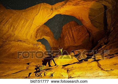 Stock Photo - "Hiker standing in illuminated rocks at night in Arches National Park, Moab, Utah, United States". Fotosearch