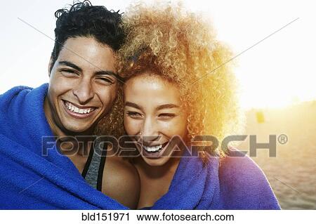 Stock Image - Hispanic couple wrapped in blanket on beach. Fotosearch