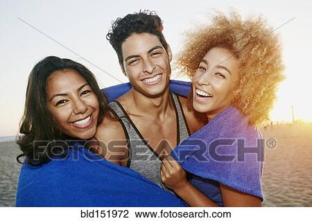 Stock Image - Hispanic friends wrapped in blanket on beach. Fotosearch