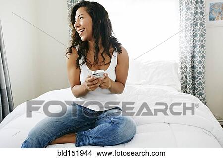 Picture - Hispanic woman using cell phone on bed. Fotosearch