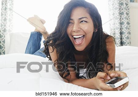 Stock Photograph - Hispanic woman using cell phone on bed. Fotosearch