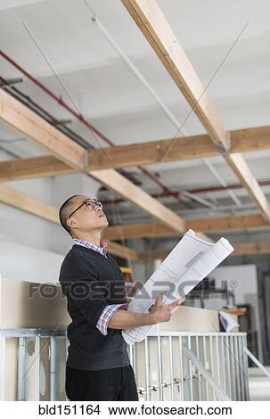 Picture - Korean architect holding blueprints and examining ceiling. Fotosearch