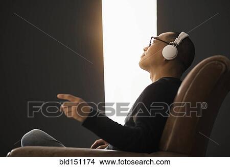 Picture - Korean man listening to headphones in armchair. Fotosearch