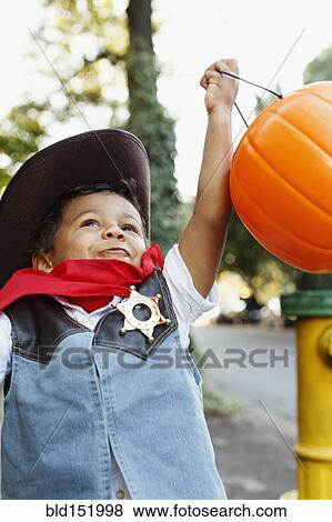 Stock Photo - Mixed race boy dressed as cowboy holding pumpkin pail for Halloween. Fotosearch