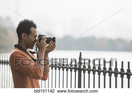 Stock Image - Mixed race man taking photograph on waterfront. Fotosearch