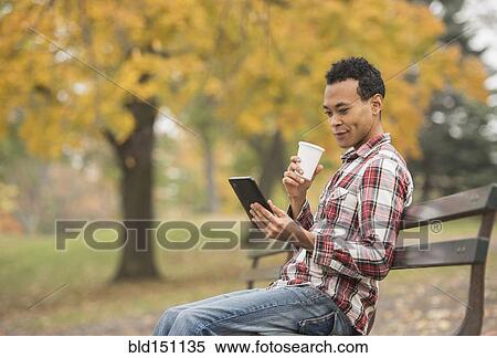 Stock Photography - Mixed race man using digital tablet on park bench. Fotosearch