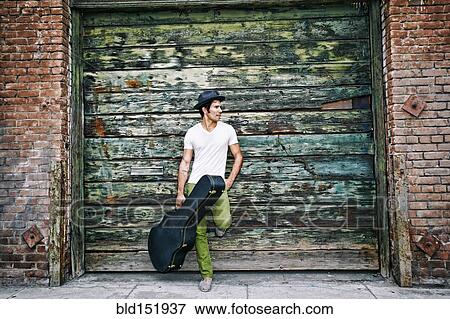 Stock Photo - Mixed race musician holding guitar case near industrial building. Fotosearch