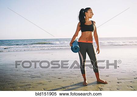 Stock Photography - Mixed race woman holding yoga mat on beach. Fotosearch
