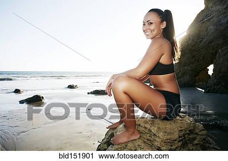 Stock Image - Mixed race woman sitting on rock on beach. Fotosearch