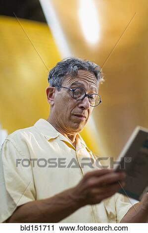 Stock Image - Older Caucasian man reading book in library. Fotosearch