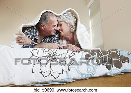 Stock Image - Older couple rubbing noses under blanket on bed. Fotosearch