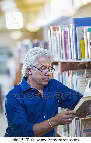 Stock Image - Older Hispanic man reading book in library. Fotosearch