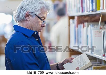 Stock Photo - Older Hispanic man reading book in library. Fotosearch