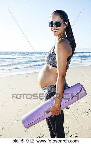 Stock Image - Pregnant Hispanic woman carrying yoga mat on beach. Fotosearch