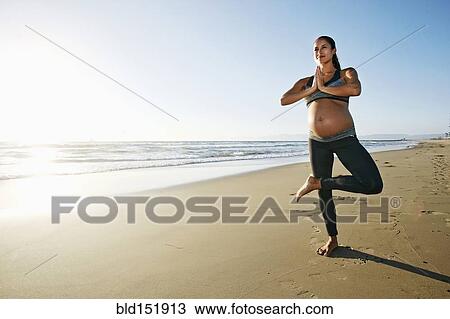 Stock Image - Pregnant Hispanic woman practicing yoga on beach. Fotosearch