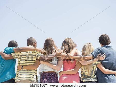 Stock Image - Rear view of teenagers hugging under blue sky. Fotosearch
