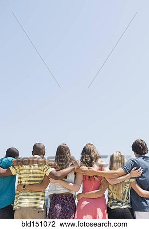 Stock Image - Rear view of teenagers hugging under blue sky. Fotosearch