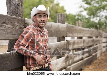 Stock Photography - Hispanic man in cowboy hat standing near fence on ranch. Fotosearch