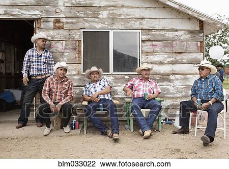 Stock Image - Hispanic men in cowboy hats relaxing near shed. Fotosearch