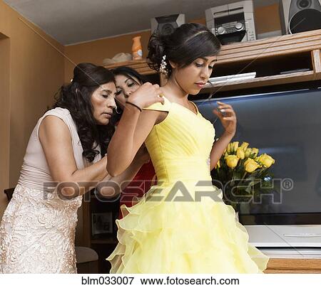 Stock Photo - Hispanic women zipping quinceanera dress in living room. Fotosearch