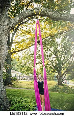 Stock Image - Acrobatic Caucasian girl hanging on fabric under tree. Fotosearch